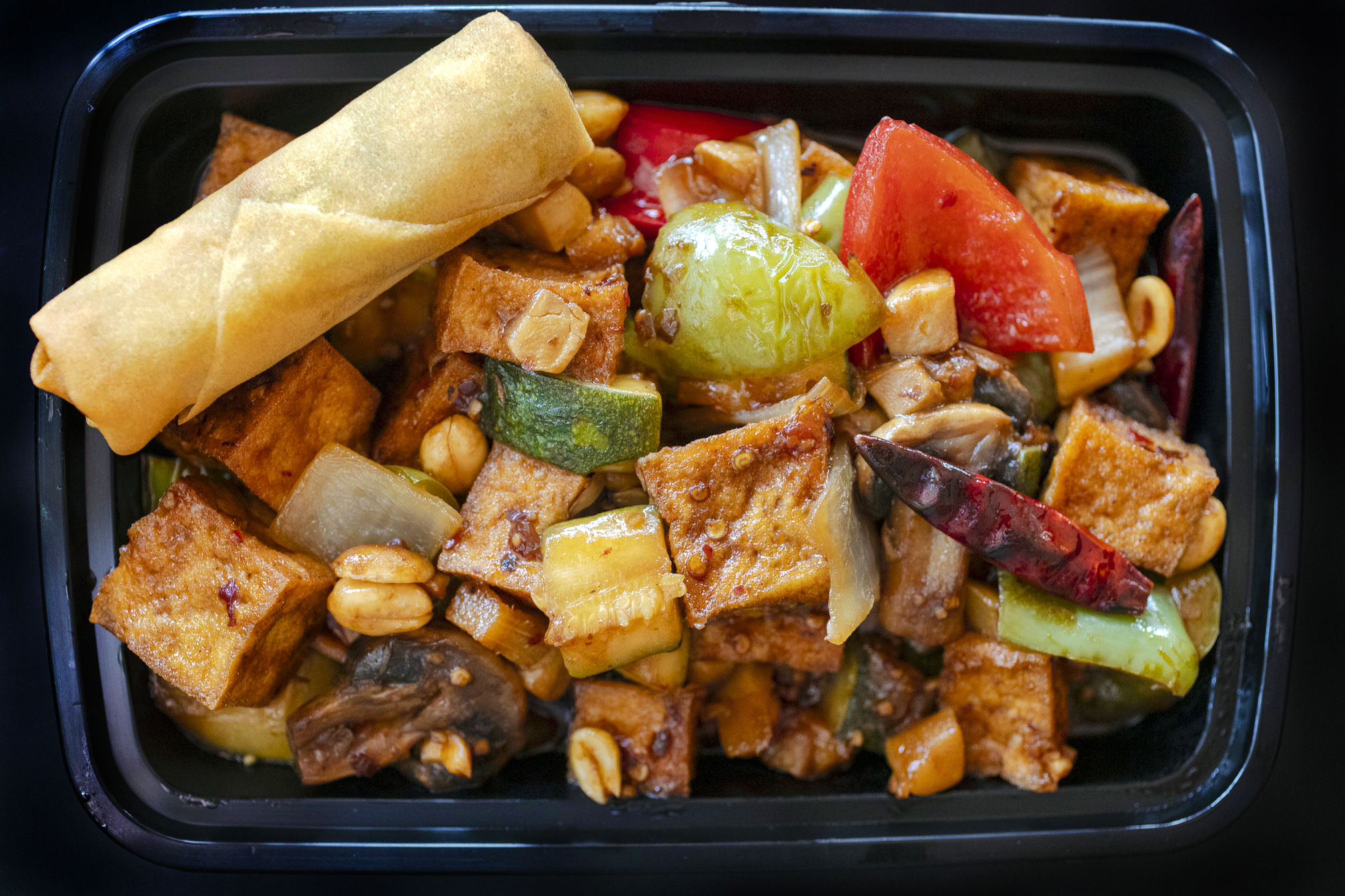 Spicy vegetarian Kung Pao Tofu served with an egg roll in a black takeout container from Asian Spice restaurant in Tucson, Arizona. Food photography by Jackie Alpers.